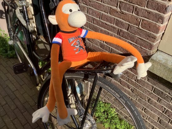 Oranje knuffel aap met lange armen, zittend op een fietsrek. Draagt een oranje shirt met "HOLLAND" erop.