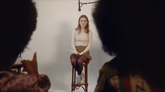 Jonge vrouw met lang bruin haar, zittend op een kruk, met een microfoon erboven. Ze draagt een crèmekleurige coltrui en een groene rok.