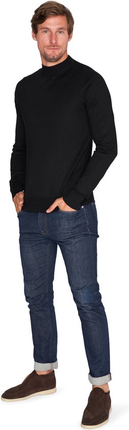 Man staat met handen in zakken, draagt zwarte coltrui en jeans.