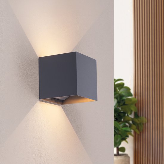 Applique murale gris foncé de forme cubique montée sur un mur clair, projetant une lumière blanc chaud vers le haut et vers le bas.