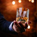 Hand met een glas amberkleurige whiskey met facet geslepen glas, achtergrond met warme, wazige lampjes.