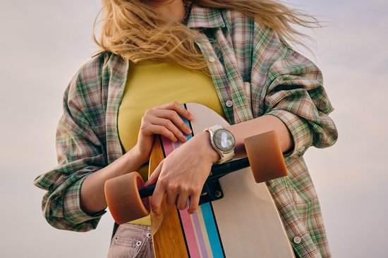Een persoon met een geel shirt, groen geruit overhemd en een witte smartwatch met oranje en blauwe accenten, die een longboard vasthoudt.