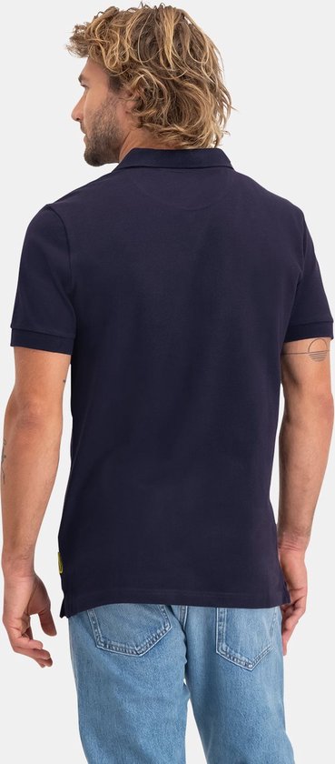 Man met kort blond haar, kijkend naar achteren, draagt een donkerblauw NOMAD poloshirt en spijkerbroek.