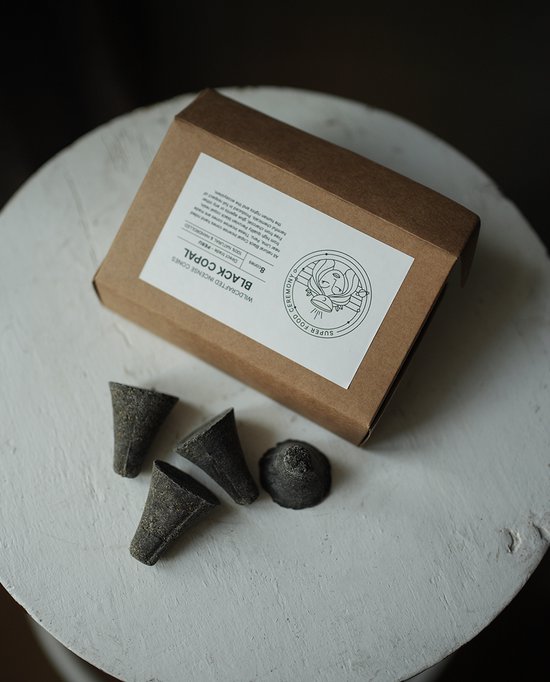 Een bruine doos met daarop 'BLACK COPAL INCENSE CONES', met vier donkergrijze, kegelvormige wierookkegels met een ruw oppervlak ernaast.