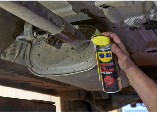 Een hand spuit WD-40 Super Kruipolie op een roestige uitlaat van een auto. De spuitbus is geel/zwart en de dop is geel.
