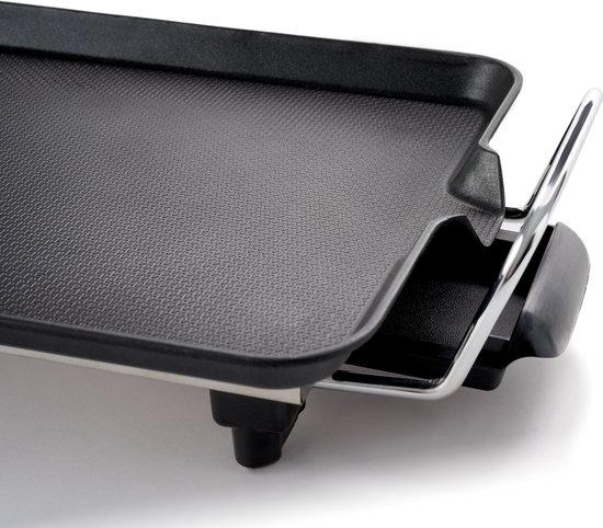 Close-up van een zwart grilloppervlak met een geribbeld gedeelte en een metalen handvat. Een deel van het chromen frame en zwarte voetjes zijn zichtbaar.