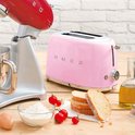 Roze SMEG broodrooster naast een rode SMEG keukenmachine op een houten tafel.