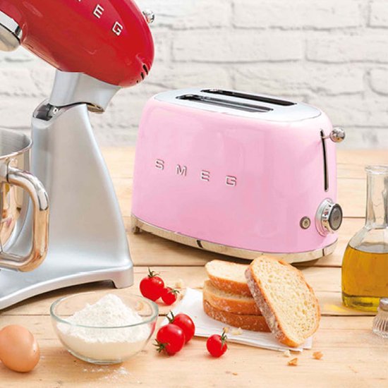 Roze SMEG broodrooster naast een rode SMEG keukenmachine op een houten tafel.