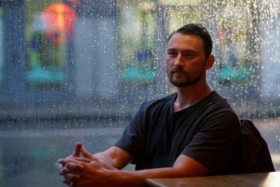 Een man met baard zit achter een tafel bij een regenachtig raam.