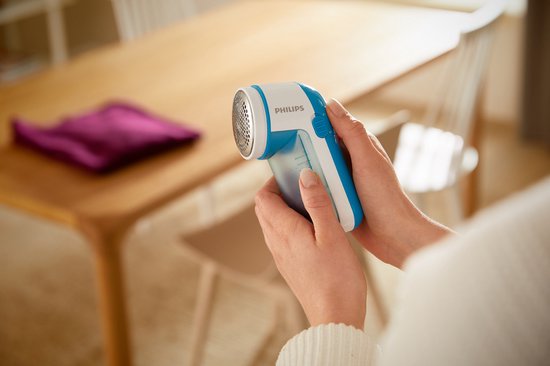 Handen die een blauwe en witte Philips pluizenverwijderaar vasthouden bij een tafel.