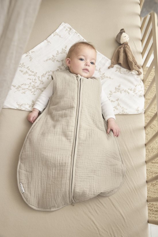 Baby in zandkleurige, gevoerde slaapzak met rits. Ligt op laken met takjesprint in een ledikant.