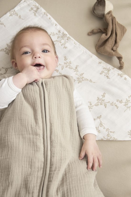 Baby lacht en sabbelt op vingers in zandkleurige slaapzak, op laken met takjesprint.