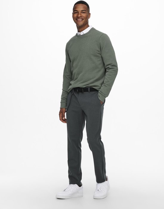 Een man met witte sneakers, een riem met ketting en een grijze broek met ruitpatroon draagt een mosgroene sweater.