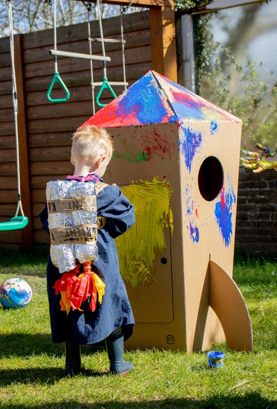 Een kind met een verfschort schildert een kartonnen raket geel, met rode en blauwe verfspatten op de punt.