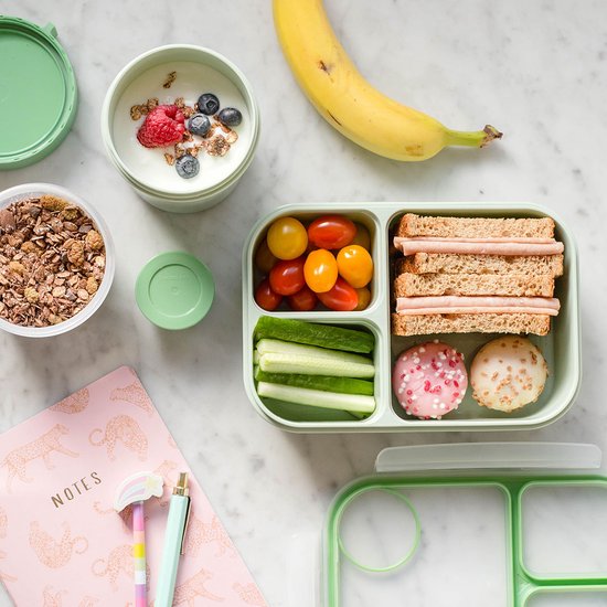 Groene lunchbox met muesli, yoghurt, fruit, groenten en een broodje.