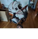 Lichtgrijze kinderwagen met koperkleurige accenten, bruine handgrepen en een zwarte babyluier.