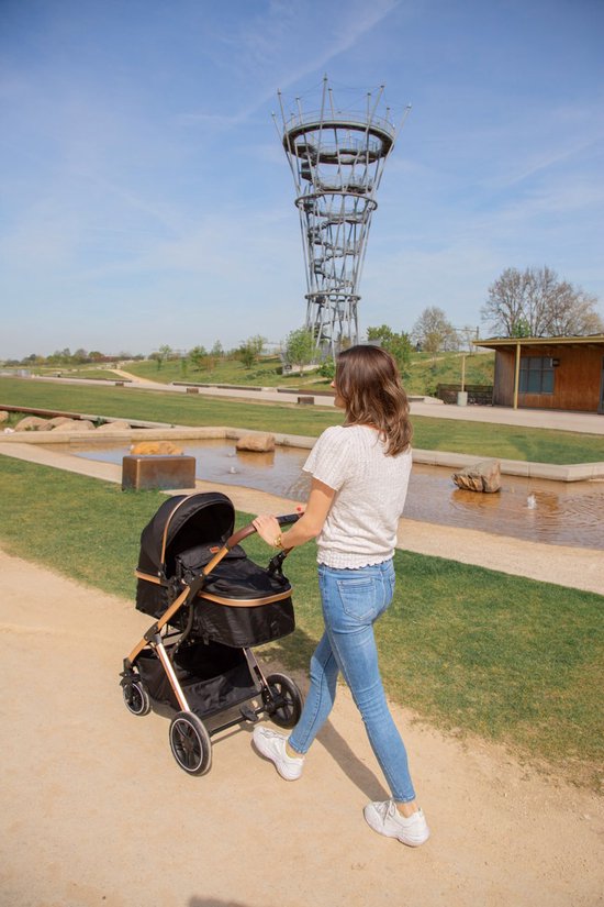 Vrouw met spijkerbroek duwt een zwarte kinderwagen met goudkleurige accenten in een park.