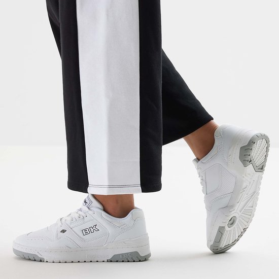 Model draagt witte lage sneakers met grijze zolen en zwarte broek met witte bies.