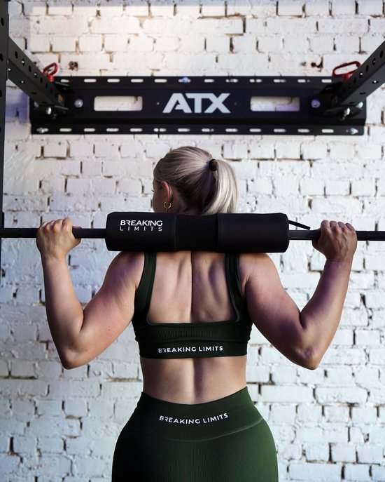 Vrouw met staartje in donkergroene sportkleding doet een squat met halterstang en zwarte pad met 'BREAKING LIMITS'.