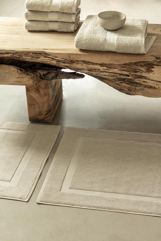Twee beige badmatten met een dubbel, ingesneden randmotief op een betonnen vloer. Twee stapels beige handdoeken en een kom liggen op een houten blad.