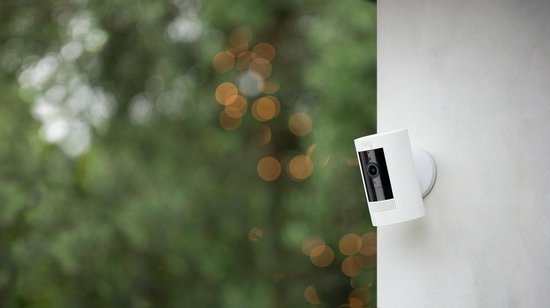 Witte Ring Stick Up Cam bevestigd aan een muur, met groene bokeh op achtergrond.