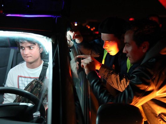 Een man met een wit T-shirt en camouflagetas zit achter het stuur, terwijl twee mannen buiten, verlicht door een zaklamp, naar hem wijzen.