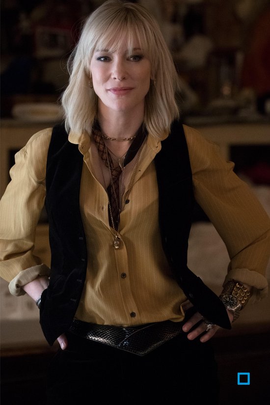 Cate Blanchett met blond haar, gekleed in een geel gestreept overhemd en zwart vest, met sieraden.