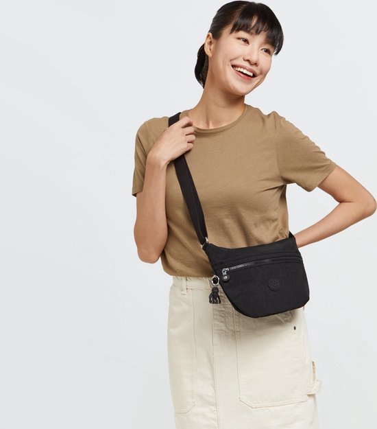 Vrouw draagt zwarte Kipling schoudertas, model Arto S, met zilverkleurige ritsen en Kipling aapje als hanger. Ze draagt een bruin T-shirt en een crème rok.