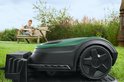 Groene en zwarte robotmaaier met een vrouw die op een bankje zit op de achtergrond.