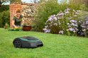 Robotmaaier op een gazon met een vrouw die bloemen plant op de achtergrond.