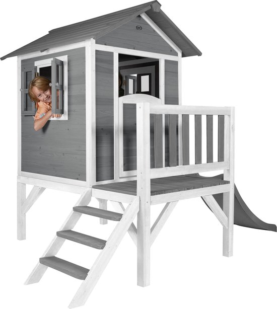 Grijs houten speelhuis met witte accenten, veranda, trap en glijbaan, met een meisje dat uit het raam kijkt.