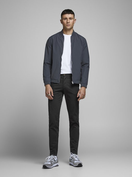 Man in donkergrijze chino broek, wit t-shirt en grijs bomberjack, gedragen met sneakers.
