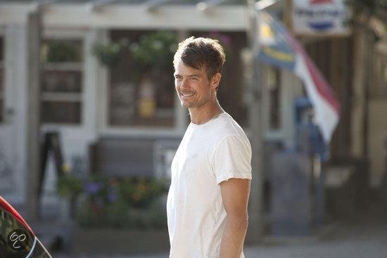Een man met kort bruin haar, een witte t-shirt, die lachend opzij kijkt.