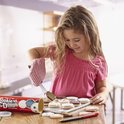 Meisje met lang blond haar, draagt roze shirt en rood-wit geblokt ovenwant. Ze gebruikt spatel om een groen-geglaceerd houten koekje van bakplaat te pakken.