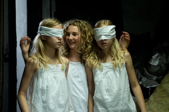 Meisje met krullen lacht terwijl twee meiden met witte blinddoeken naast haar staan.