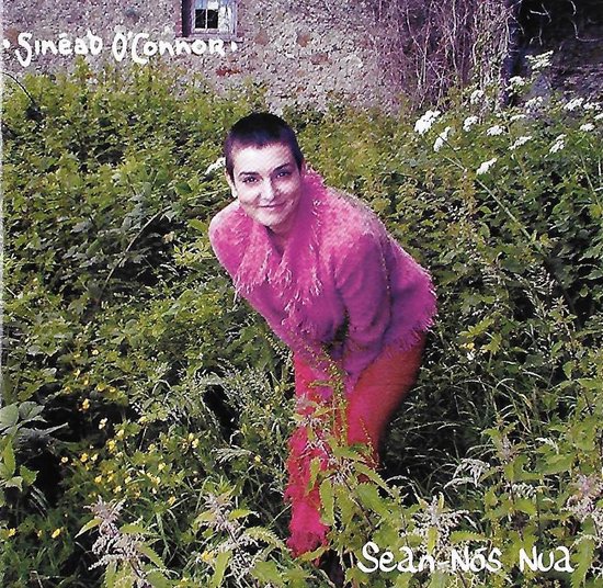 Sinead O'Connor in een roze trui en rode broek, omringd door groen gras en bloemen. Tekst: Sean-Nos Nua.
