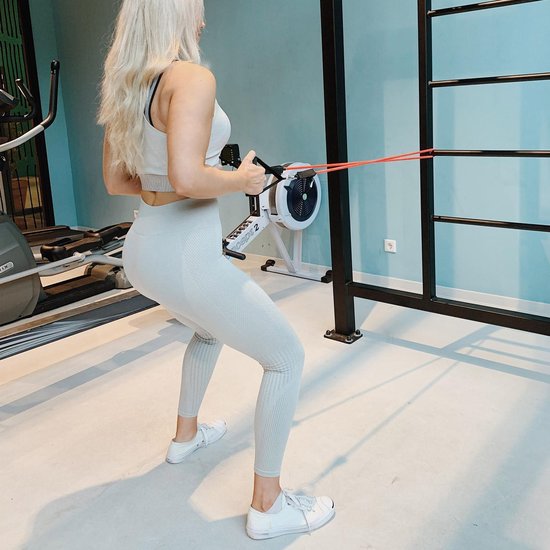 Vrouw met blond haar in lichte sportkleding doet een squat met een rode weerstandsband om haar voeten.