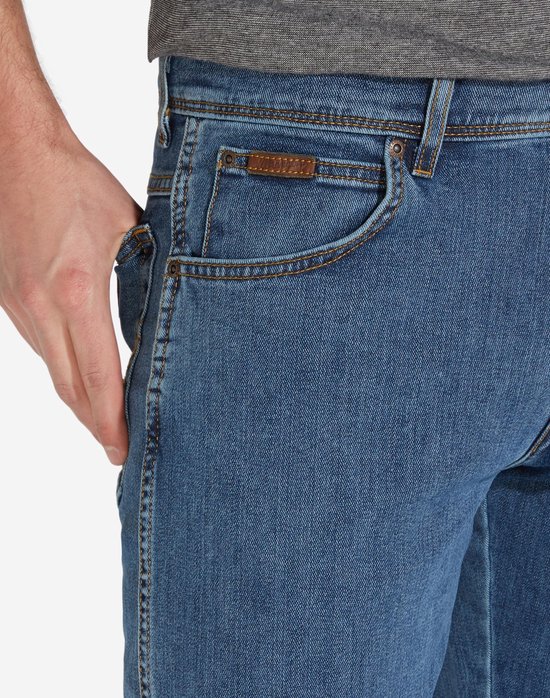 Close-up van de voorkant van een blauwe spijkerbroek met bruin lederen Wrangler logo op de zak.