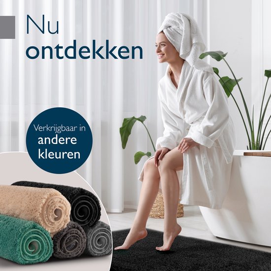 Een vrouw in een badjas zit op de rand van een bad, naast een stapel opgerolde badmatten in beige, groen en grijs. Een donkergrijze badmat ligt op de vloer.