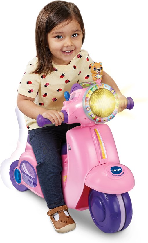 Meisje zit op een roze VTech loopscooter met paarse details, knipperende lichten en een poppetje.
