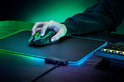 Hand die een zwarte Razer Basilisk V3 X HyperSpeed gamingmuis met groen scrollwiel vasthoudt op een zwart muismat met RGB-rand.