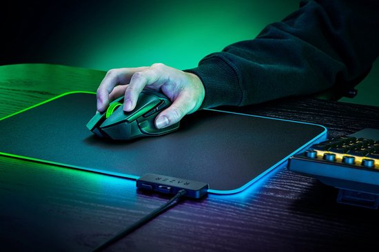 Hand die een zwarte Razer Basilisk V3 X HyperSpeed gamingmuis met groen scrollwiel vasthoudt op een zwart muismat met RGB-rand.