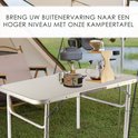 Zilverkleurige campingtafel met kampeerspullen zoals een brander, mokken en een waterkoker, in een buitenomgeving.