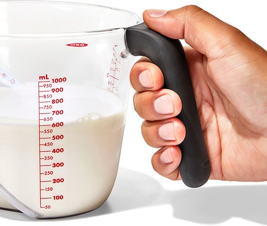 Une main tient la poignée noire et ergonomique d'un pichet mesureur transparent avec un liquide blanc.