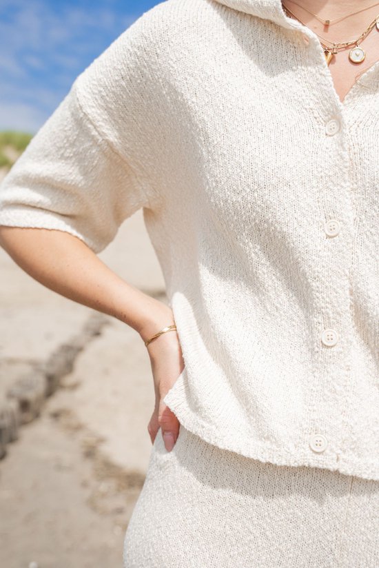 Off-white gebreide top met korte mouwen en knoopsluiting, gedragen door een model op het strand.