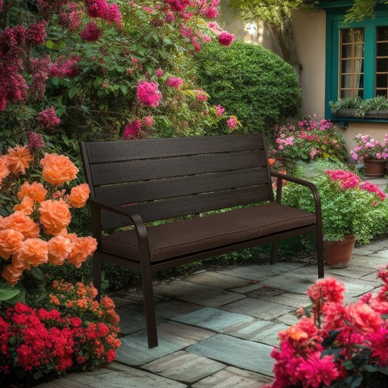 Donkerbruine tuinbank met kussen in een tuin vol bloemen.