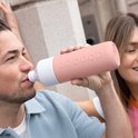 Man drinkt uit Dopper Insulated drinkfles in pebble peach, met een vrouw op de achtergrond.