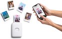 Handen houden een smartphone vast met een Instax foto-app. Naast de smartphone een witte Instax Mini Link 2 printer, met een foto van twee personen met een hartje erop.