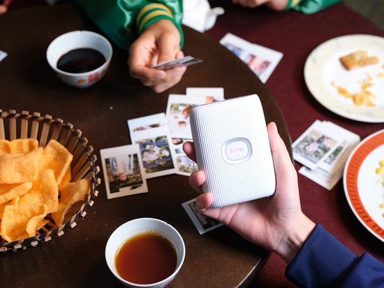 Hand houdt een witte Instax Mini Link 2 printer vast boven een tafel met prints en snacks. Twee mensen aan tafel met Instax foto's.