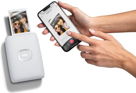 Handen houden een smartphone vast met een Instax foto-app. Naast de smartphone een witte Instax Mini Link 2 printer, met een foto van twee lachende personen die naar de camera kijken.